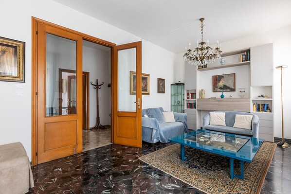 Una sala de estar con un sofá azul, una mesa de centro de vidrio, una araña y una alfombra estampada.