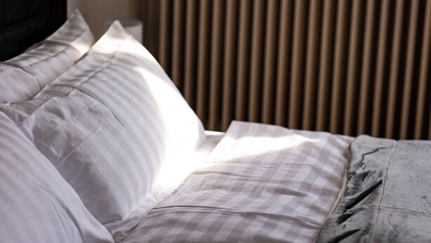 A neatly made bed with white and grey bedding, a black headboard, and vertical blinds in the background. - ME LIVING 1: Hauptallee - Bad Pyrmont - Apartment (Bad Pyrmont)