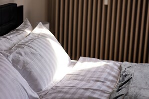 Una cama bien hecha con ropa de cama blanca y gris, un cabecero negro y persianas verticales al fondo.