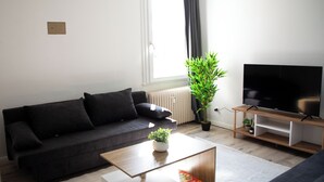 A modern living room with a black sofa, a wooden coffee table, a flat-screen TV on a wooden stand, a potted plant, and a radiator. - ME LIVING 1: Hauptallee - Bad Pyrmont - Apartment (Bad Pyrmont)