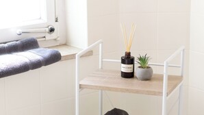 A bathroom with a white tiled wall, a wooden shelf with towels and a hairdryer, a small potted plant, and a bottle of reed diffuser. - ME LIVING 1: Hauptallee - Bad Pyrmont - Apartment (Bad Pyrmont)