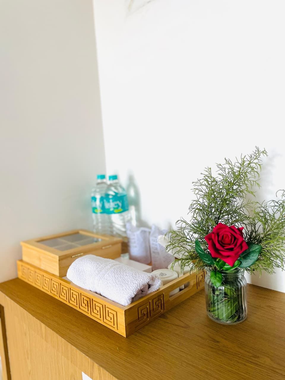 A wooden tray with a white towel, a glass vase with a red rose, and water bottles on a wooden surface.