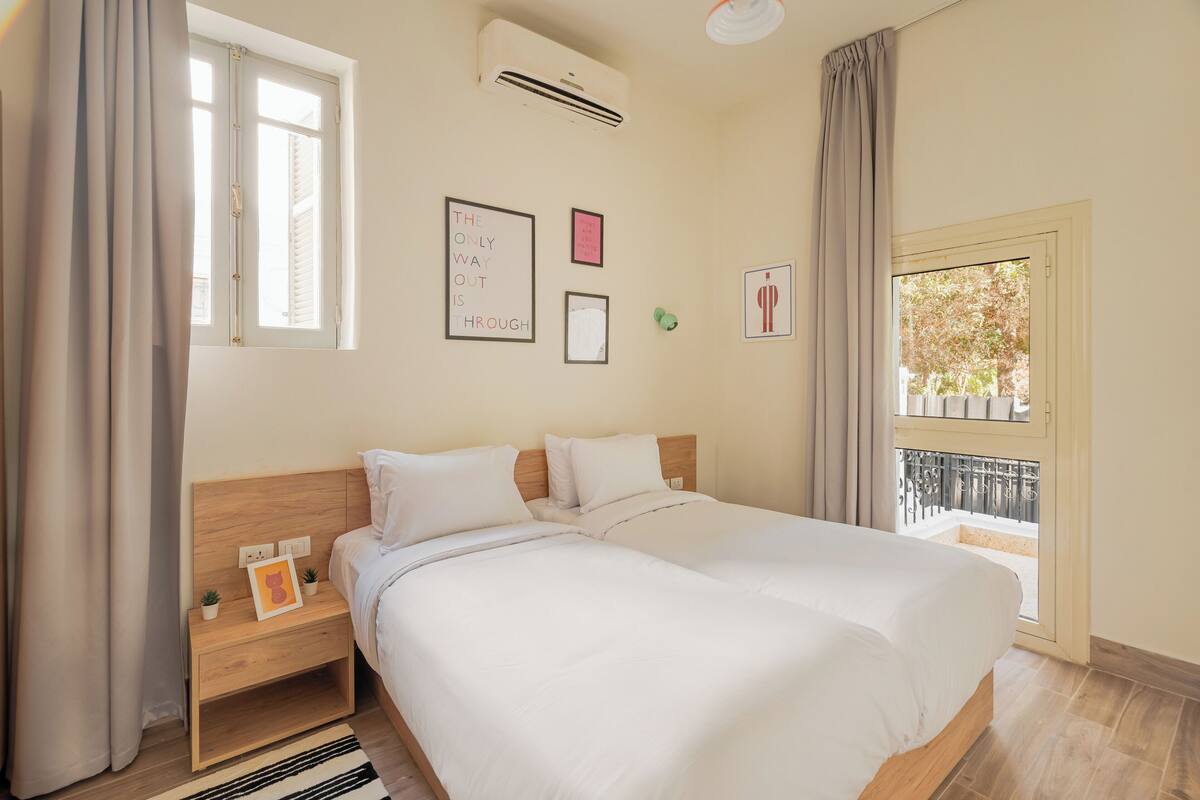 A bedroom with a double bed, a nightstand, a wall-mounted air conditioner, and a window with curtains.