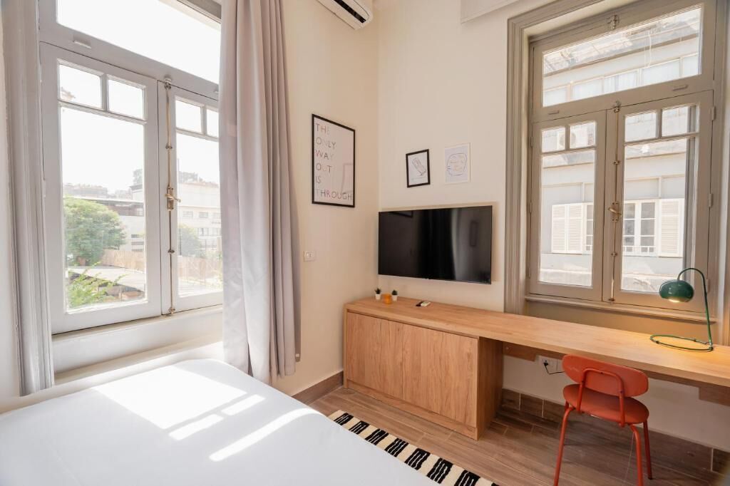 Un dormitorio con cama, escritorio, silla, televisor y ventanas grandes.
