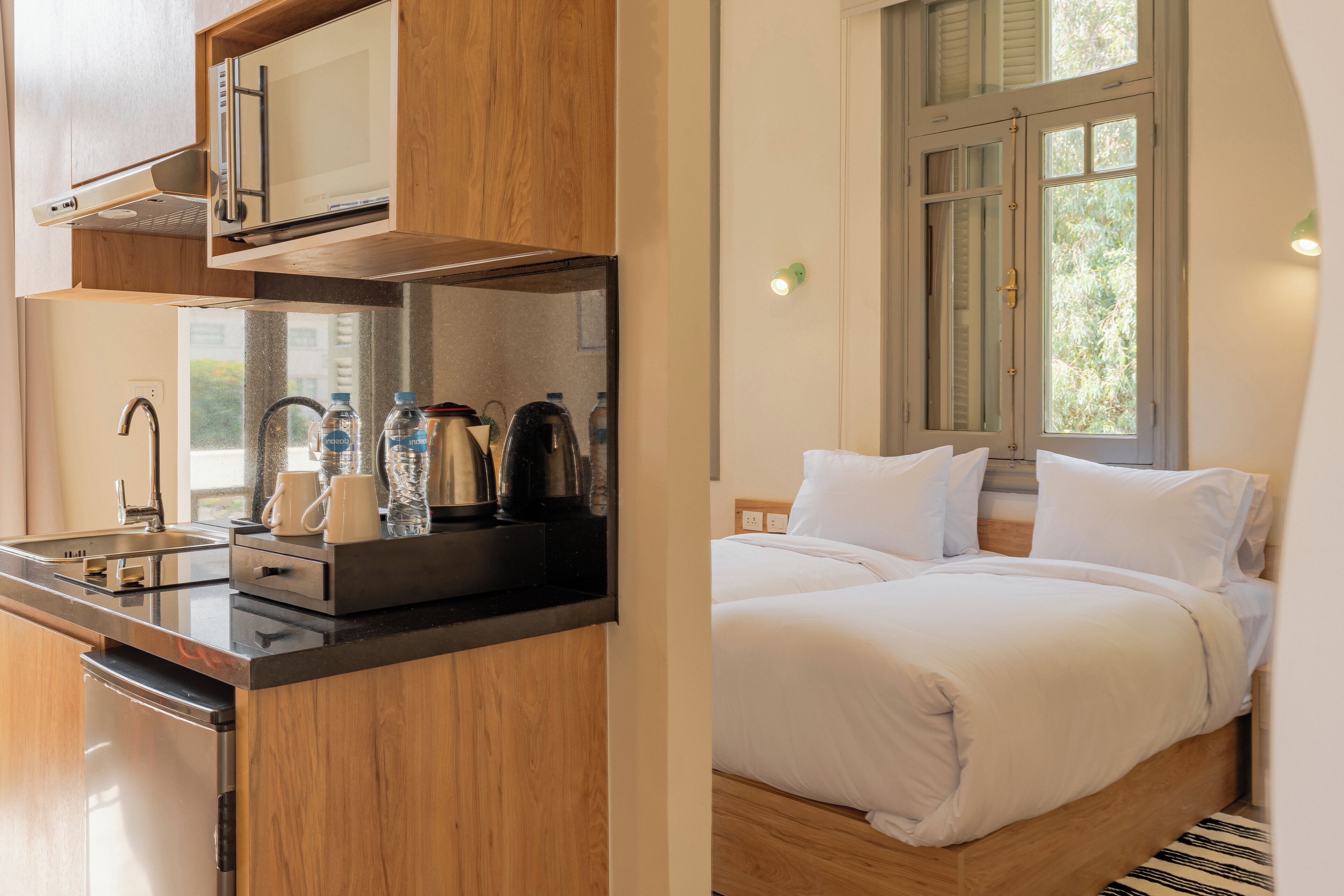 A modern hotel room with a kitchen area, a bed with white linens, and a window with a view of greenery.