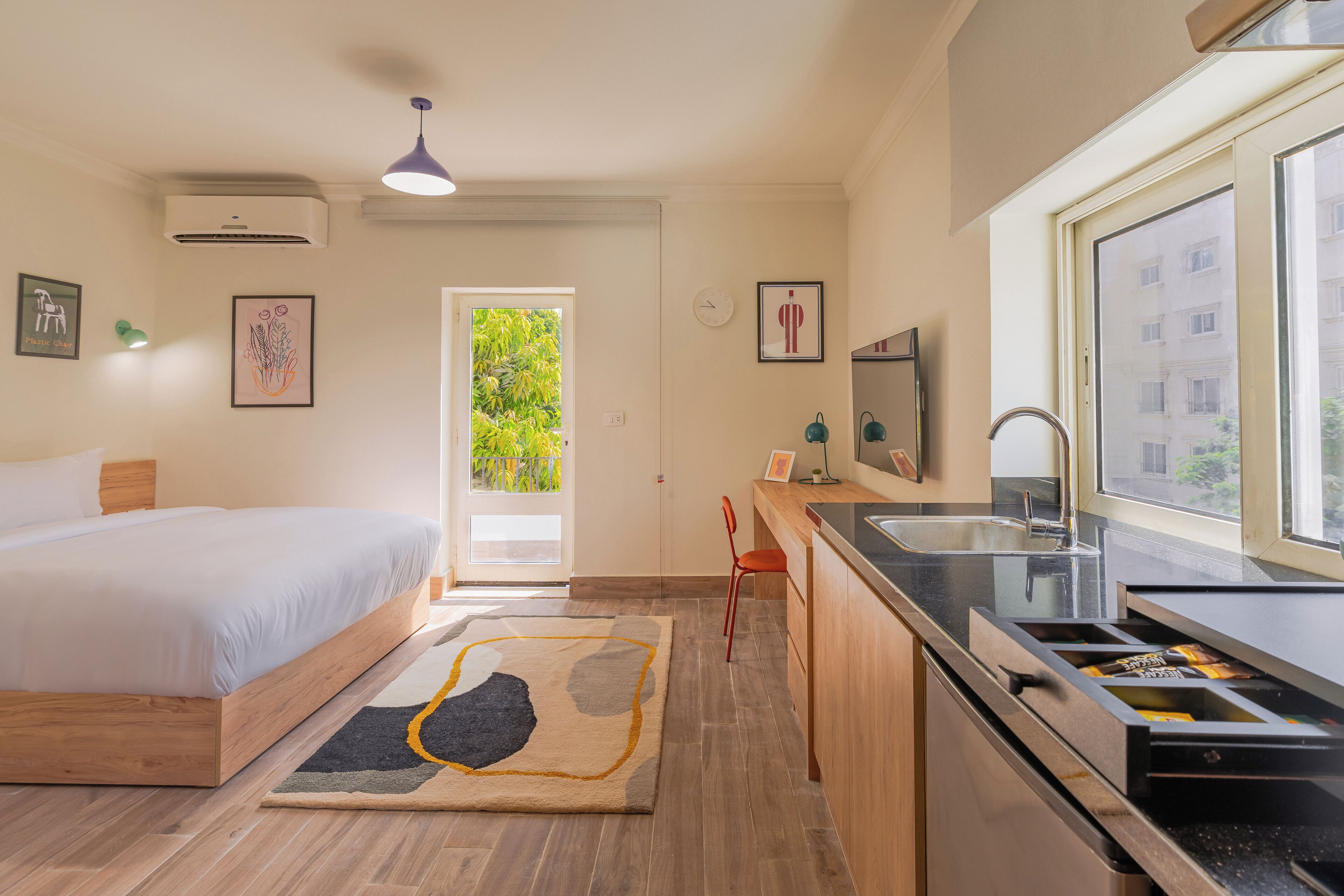 A modern bedroom with a bed, a desk, and a kitchen area with a sink and window.
