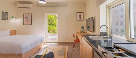 A modern bedroom with a bed, a desk, and a kitchen area with a sink and window.