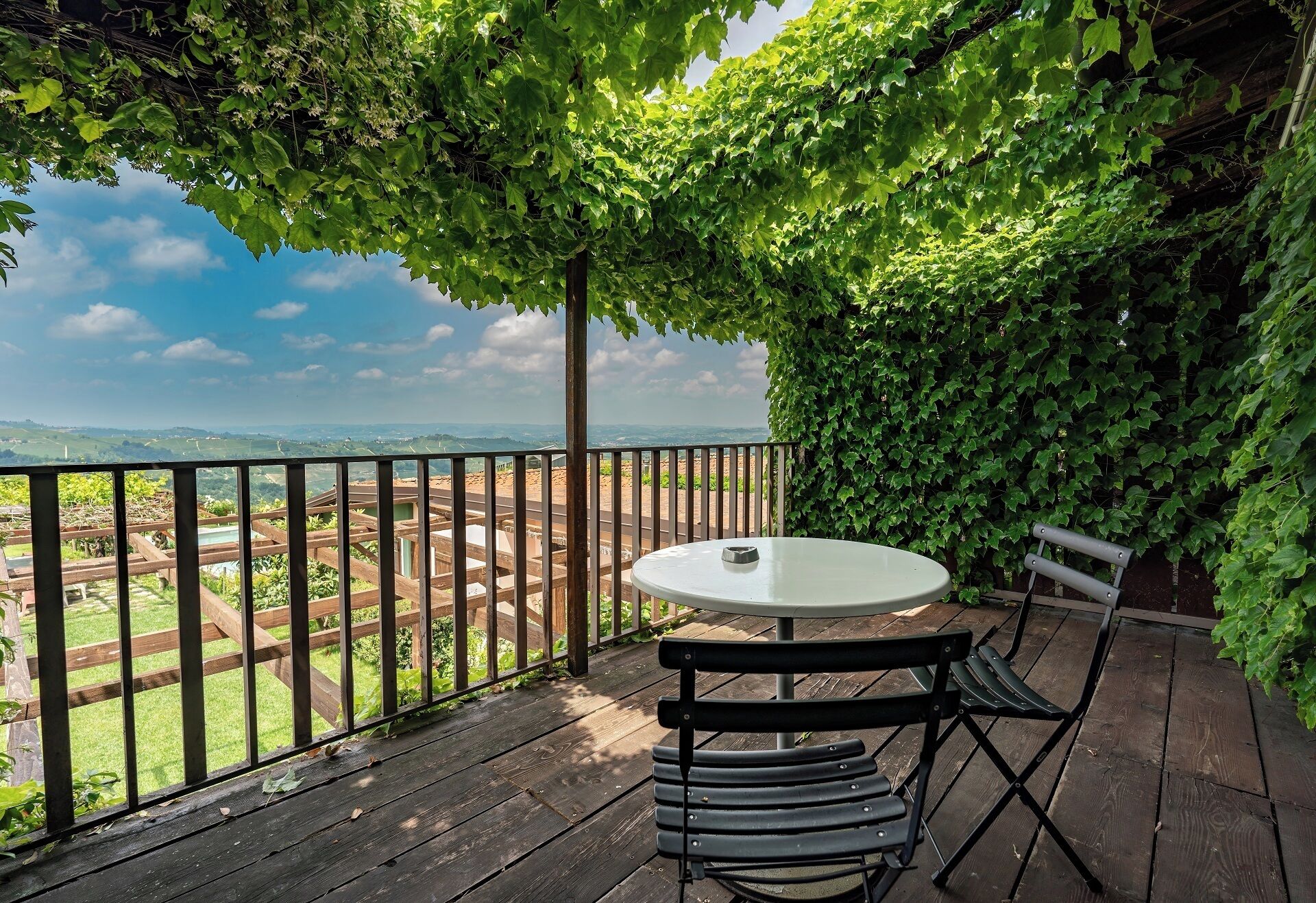 A wooden deck with a round table and two chairs, surrounded by a metal railing and lush green foliage.