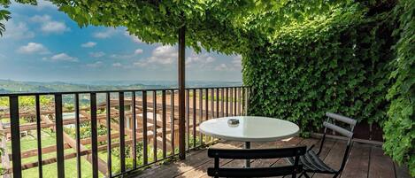 A wooden deck with a round table and two chairs, surrounded by a metal railing and lush green foliage.