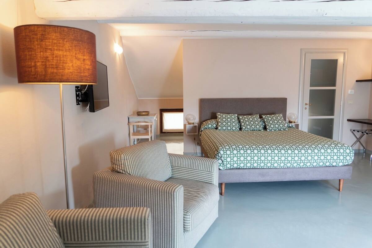 Une chambre à coucher avec un lit, deux fauteuils à rayures, une lampe sur pied, une petite table avec un bol, et un téléviseur fixé au mur.