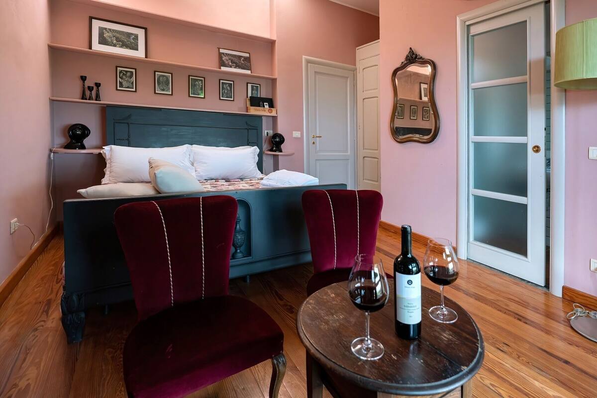 Une chambre à coucher avec un lit, deux chaises, une table avec des verres à vin et une bouteille, et un miroir au mur.