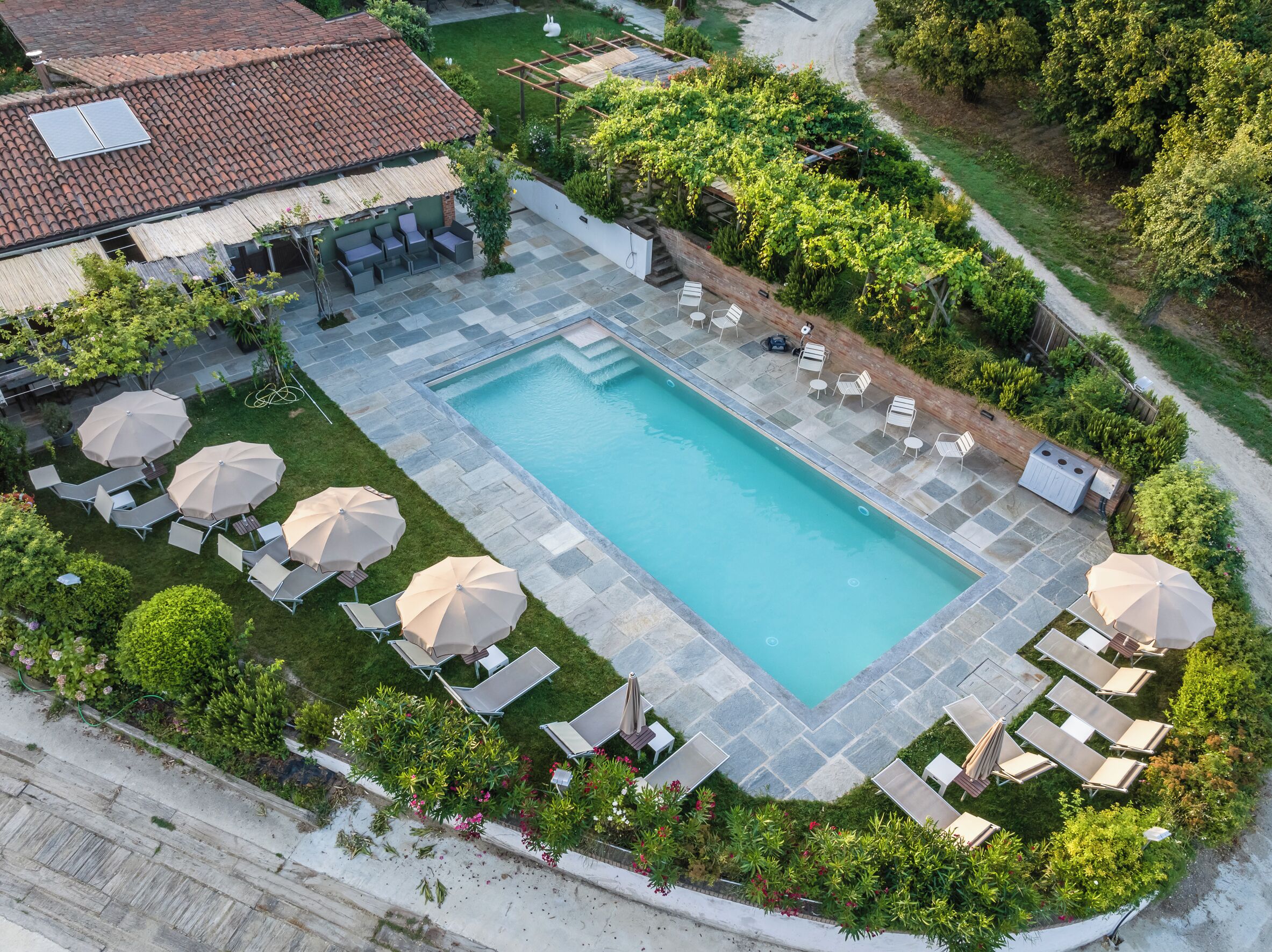An aerial view of a swimming pool with lounge chairs and umbrellas, surrounded by greenery and a building with a tiled roof.