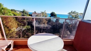 Un balcon avec une table ronde et des chaises blanches, donnant sur une zone côtière bordée d’arbres et de bâtiments.