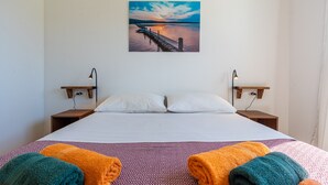 Ein Hotelzimmer mit einem Bett, zwei Nachttischen, einem Wandbild mit Sonnenuntergang über Wasser und zwei zusammengerollten Handtüchern auf dem Bett.