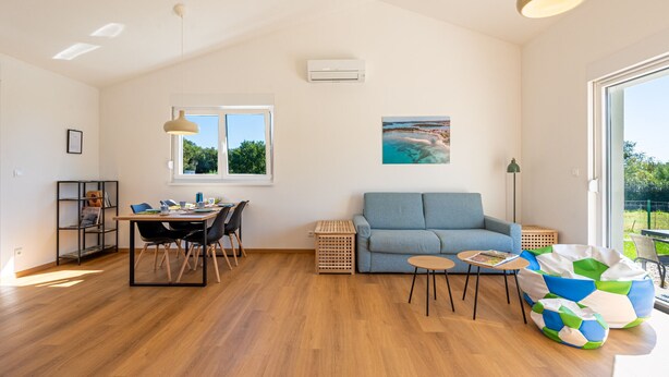Ein geräumiges Wohnzimmer mit Holzboden, einem blauen Sofa, einem Esstisch mit Stühlen, einem kleinen Couchtisch, einem bunten Sitzsack, einem Bild an der Wand und einem Fenster mit Tageslicht.