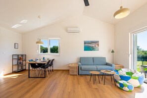 A spacious living room with a wooden floor, a blue sofa, a dining table with chairs, a small coffee table, a colorful bean bag, a painting on the wall, and a window providing natural light. - Renata’s Pool House Medulin (2+2) (Medulin)