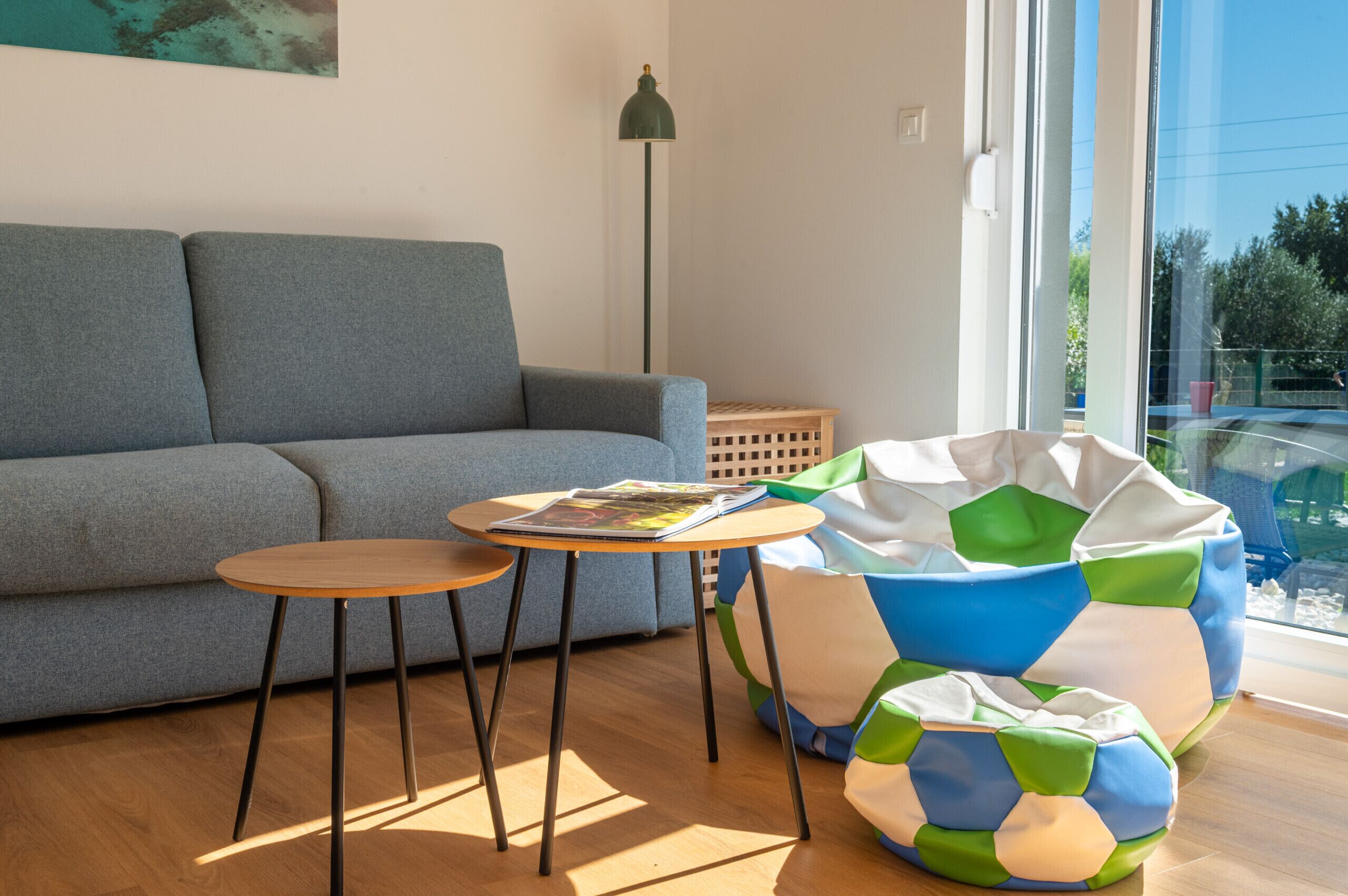 Un salon moderne avec un canapé gris, une table ronde en bois sur laquelle repose un magazine, un lampadaire et un fauteuil en forme de poire coloré.