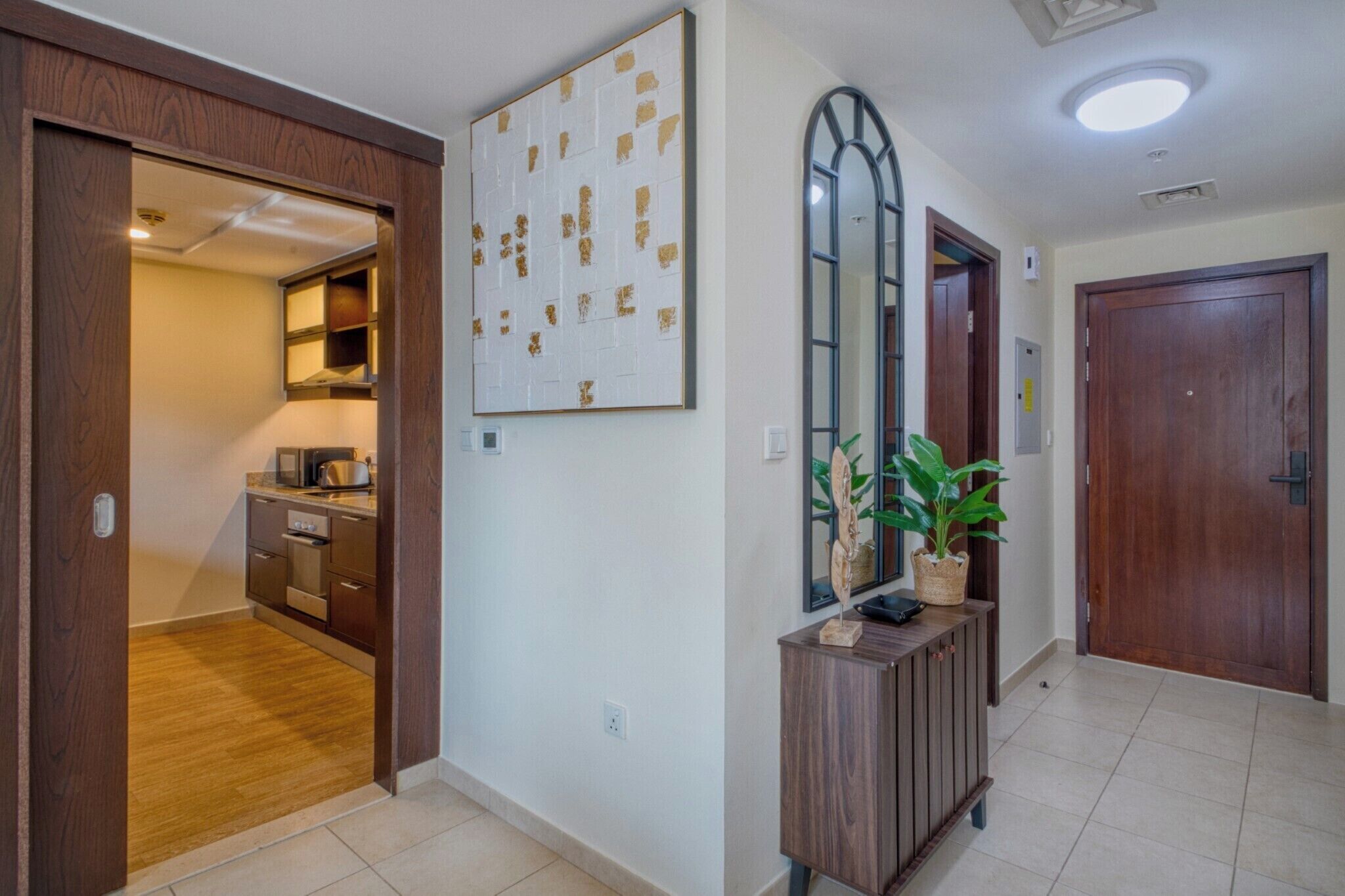 Un couloir avec une porte en bois, un miroir, une œuvre d’art accrochée au mur, une plante en pot et un coin cuisine visible à travers une porte ouverte.
