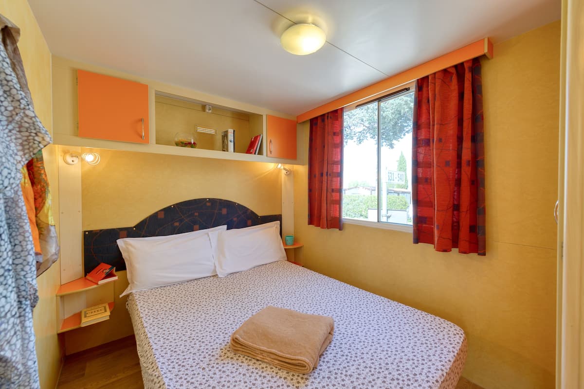 A bedroom with a bed, a nightstand, a shelf with books, a window with curtains, and a ceiling light.