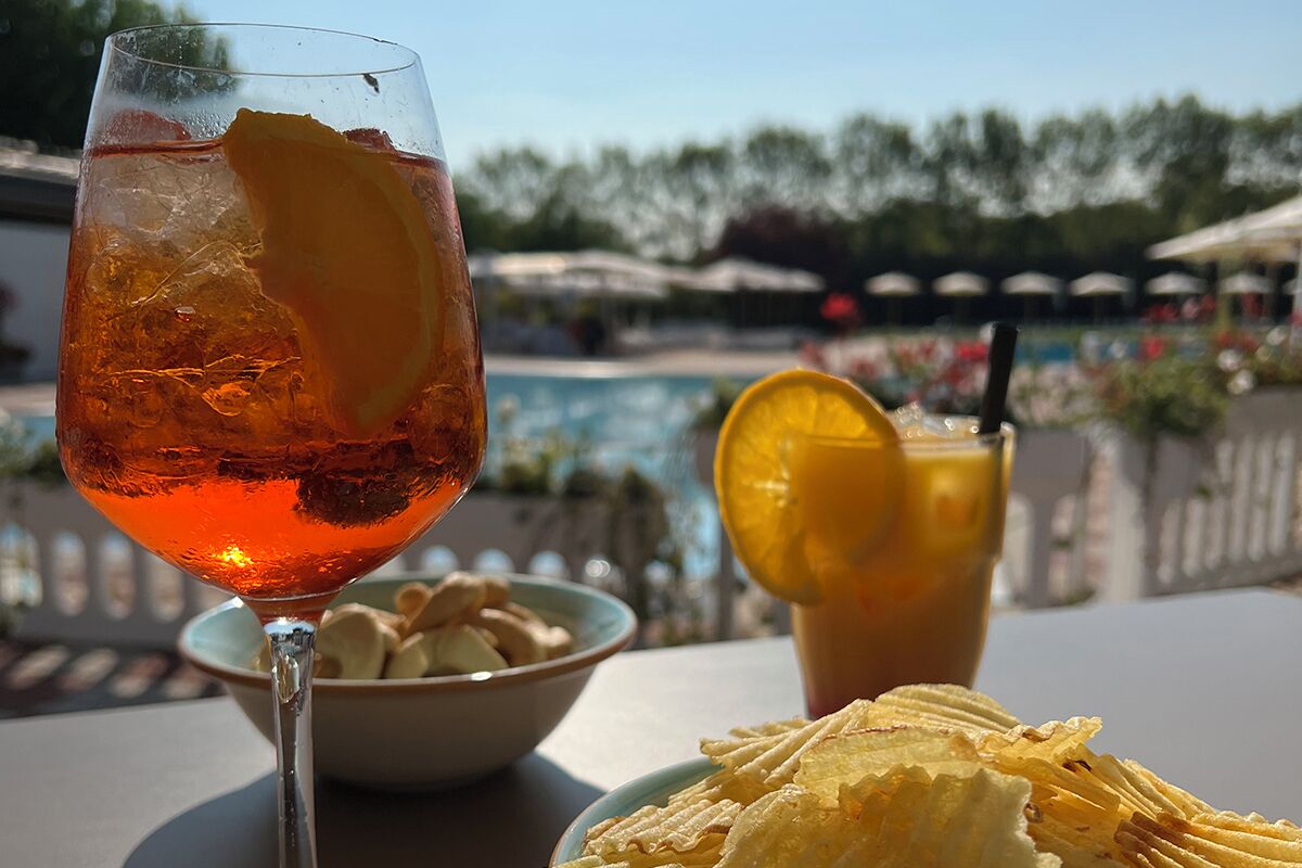 Un vaso de bebida color ámbar con hielo y una rodaja de limón, un cuenco de galletas y un plato de papas fritas sobre una mesa, con una piscina y sombrillas al fondo.