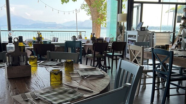 A restaurant with outdoor seating overlooking a lake, featuring wooden tables, chairs, and a view of mountains.