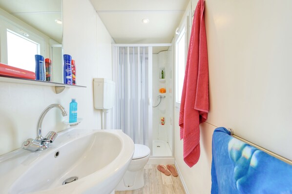 Un baño moderno con lavamanos, inodoro y ducha blancos. Hay artículos de aseo en la repisa, y una toalla roja cuelga en la pared.