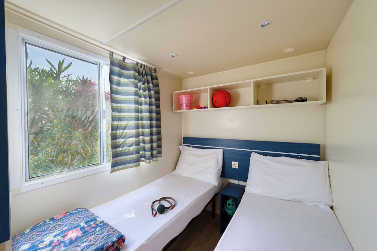 A small, single-bed room with a window, a curtain, a shelf with a red ball, and a bucket.