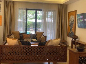A living room with a sofa, a coffee table, and a laptop. There is a window with curtains, a painting on the wall, and a wooden partition. - Nana in Diani’s  cozy 1bed luxury apartment w/beach,pool access! (Kwale County)