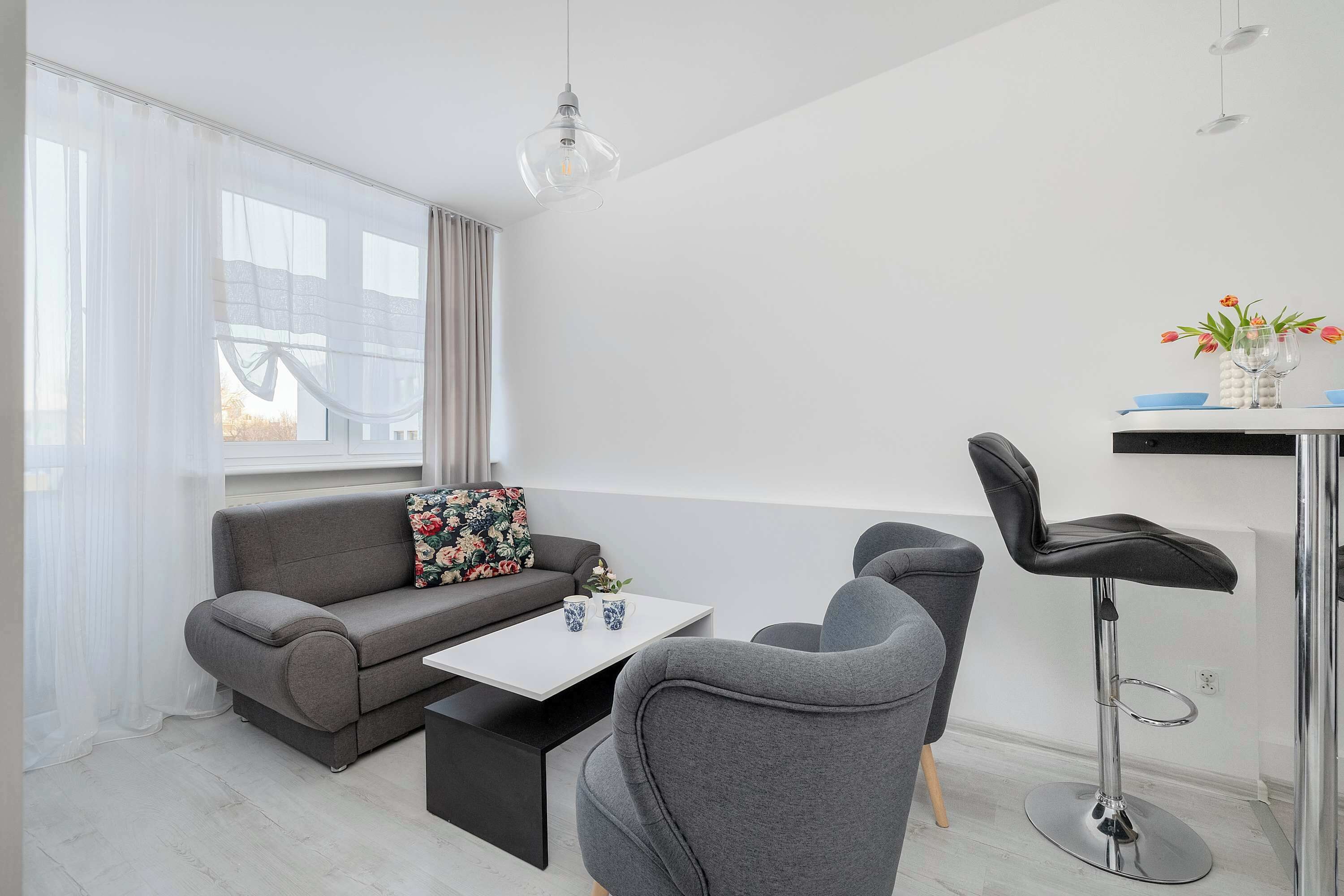 A modern living room with a grey sofa, a coffee table, a bar stool, and a window with sheer curtains.