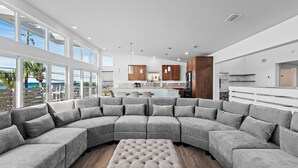 A modern living room with a large grey sectional sofa, a tufted ottoman, and a view of the kitchen and outdoor seating area through large windows. - 6BR Gulf-Front w/ Private Beach & Pool (Panama City Beach)