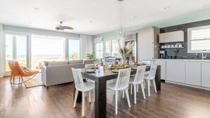 A modern kitchen with white cabinets, a dining area with a dark wooden table, and a view of the beach through large windows. - High Tide | Oceanfront 3BR with Pool & Beach Access | Ocean Point (Isle of Palms)