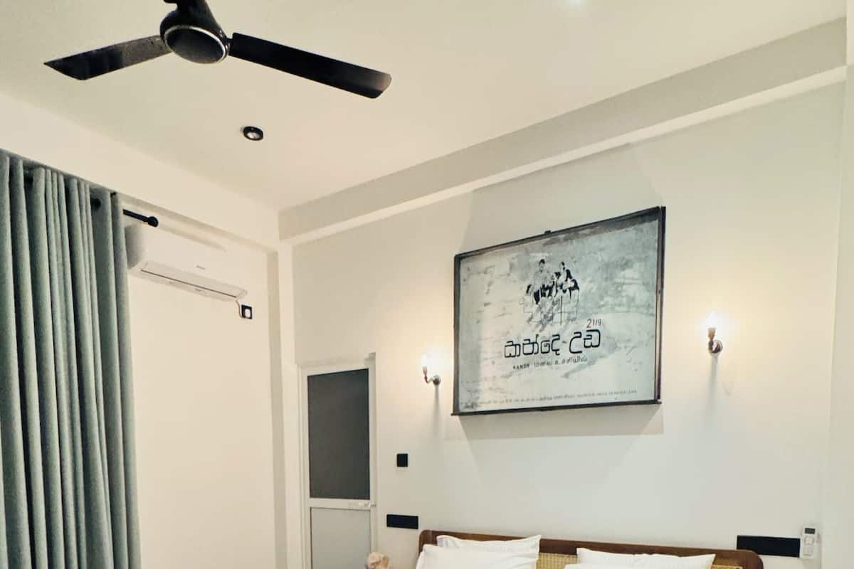 A hotel room with a bed, a ceiling fan, a wall-mounted air conditioner, a framed picture, and a nightstand with a lamp.