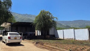Une voiture blanche est garée devant un bâtiment doté d’un porche abrité, avec un chemin de terre et une colline en arrière-plan.