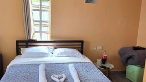 Un lit bien fait, avec des serviettes à portée de main, une fenêtre avec des rideaux, une petite table sur laquelle se trouvent une bouteille et une boîte de mouchoirs, et un panier vert posé sur le sol.