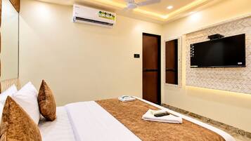 A hotel room with a bed, pillows, a remote control, a TV, and an air conditioning unit.