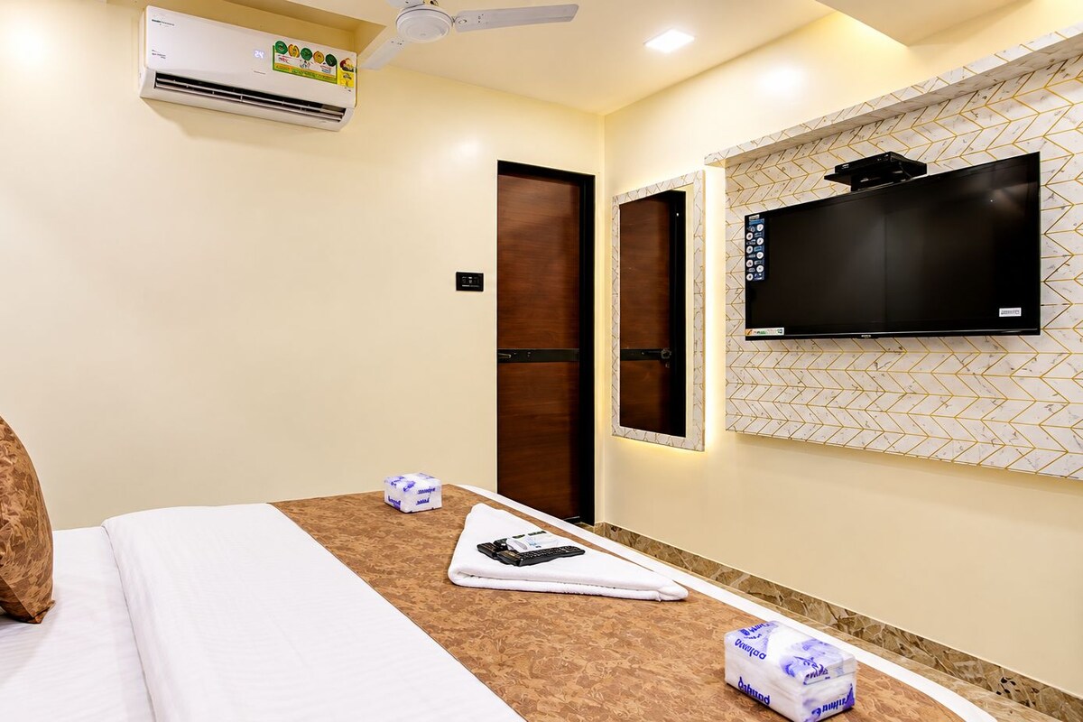 A hotel room with a bed, pillows, a flat-screen TV mounted on the wall, an air conditioning unit, and a door.