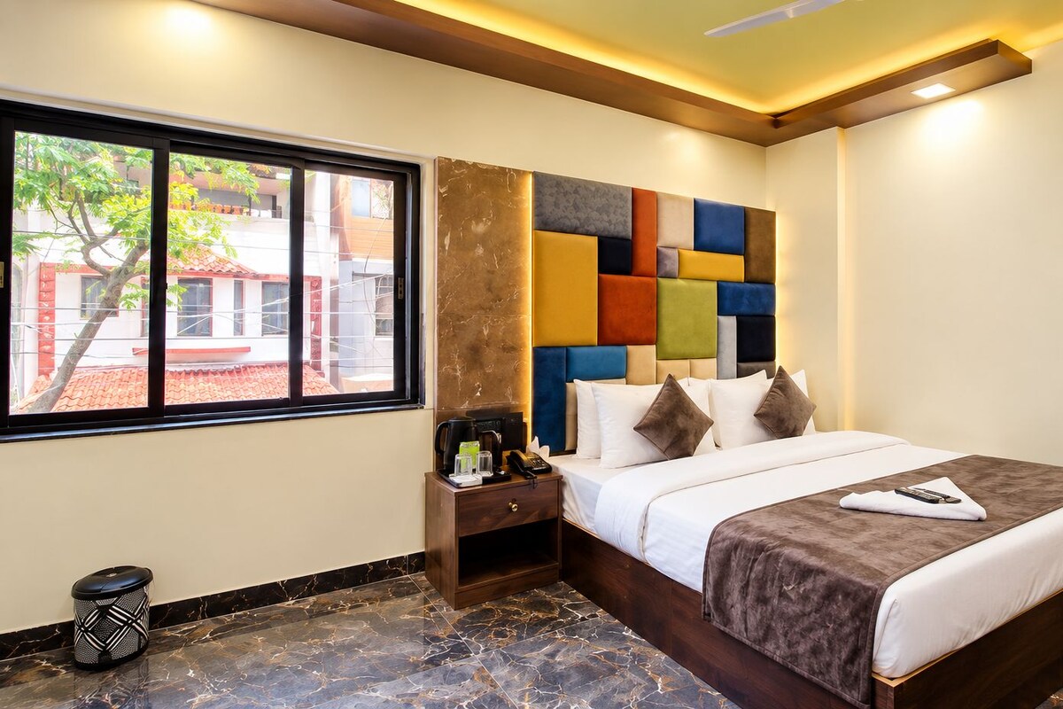 A hotel room with a bed, bedside table, and a window with a view of buildings.