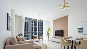 Una sala de estar moderna con sofá, mesa de comedor y sillas. Hay un televisor montado en la pared y un gran ventanal con cortinas.