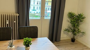 Ett rum med ett träbord, två stolar, en krukväxt och en utsikt över byggnader genom ett fönster med gardiner.