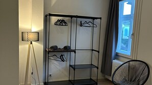 Ett rum med en svart metallhylla där kläder hänger, en golvlampa, en rund stol och ett fönster med gardiner.