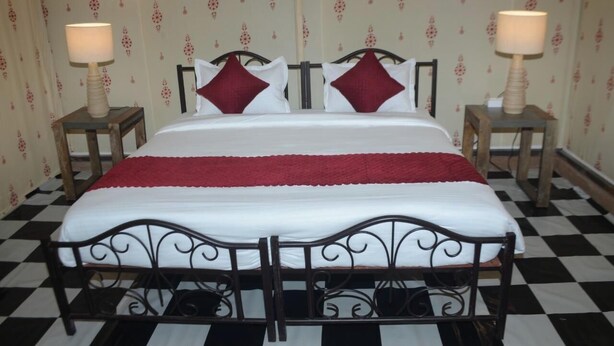 A hotel room with a checkered floor, a bed with a white and red bedspread, two nightstands with lamps, and patterned wallpaper. - Pushkar Dhani Camps And Resort (Pushkar)