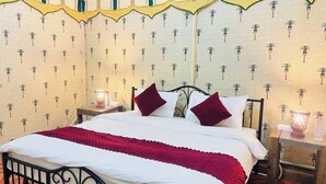 A bed with white bedding and a red blanket, two red pillows, a nightstand with a lamp, and a wall with palm tree patterns. - Pushkar Dhani Camps And Resort (Pushkar)