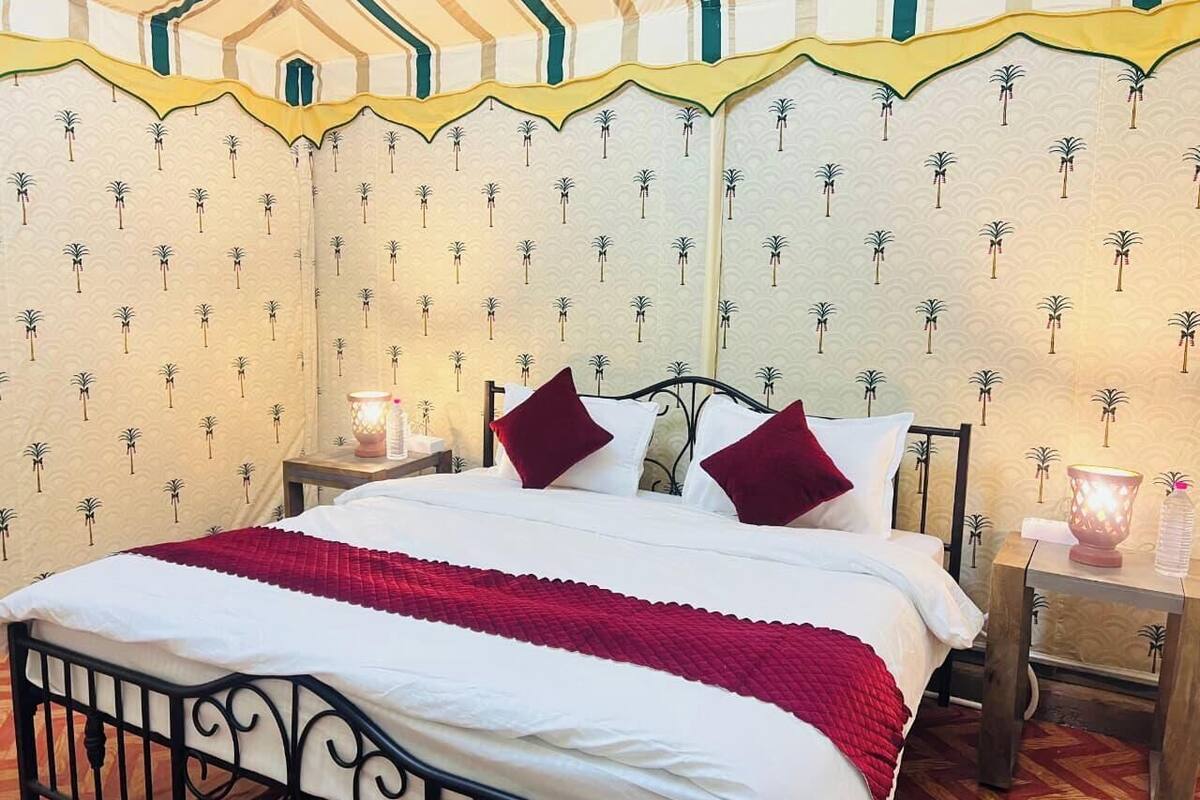 Un lit avec une literie blanche et une couverture rouge, deux oreillers rouges, une table de chevet avec une lampe, et un mur orné de motifs de palmiers.
