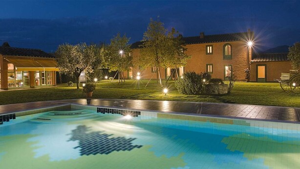 A swimming pool with a tiled deck, a covered seating area, and a building with multiple windows and a door.