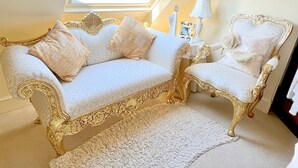 Una habitación lujosa con un sofá blanco y un sillón a juego, ambos con detalles dorados. Un tapete blanco cubre el suelo, y hay una mesita con una lámpara y fotos enmarcadas.