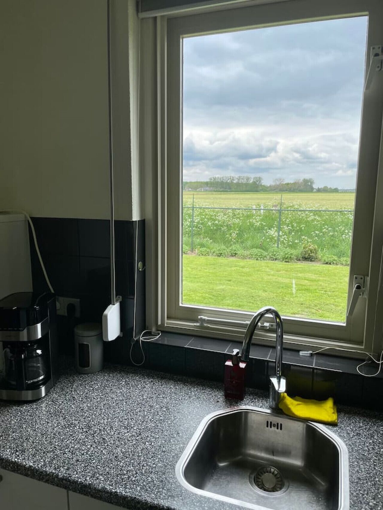 A kitchen with a stainless steel sink, a coffee maker, and a window overlooking a green field.