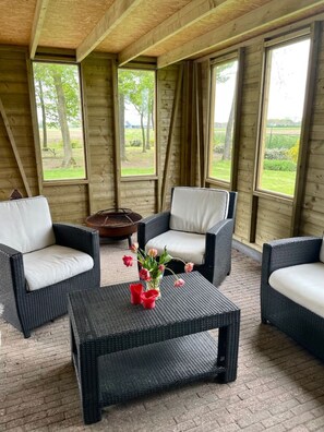 Een gezellig buiten zitgedeelte met rieten meubels en een centrale tafel, grote ramen met uitzicht op een groen landschap.