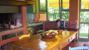 Una mesa de madera con sillas, un frutero y una cocina con estufa y lavaplatos.