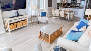 Una sala de estar moderna con sofá, mesa de centro y televisor. Se ve un comedor con sillas y una cocina al fondo.