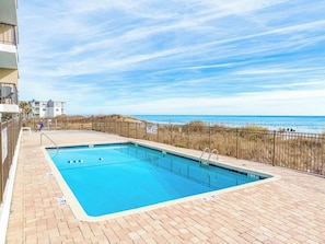 A rectangular swimming pool with a tiled edge, surrounded by a brick deck, and a view of the beach and ocean in the background. - Sea Marsh I 404 (North Myrtle Beach)